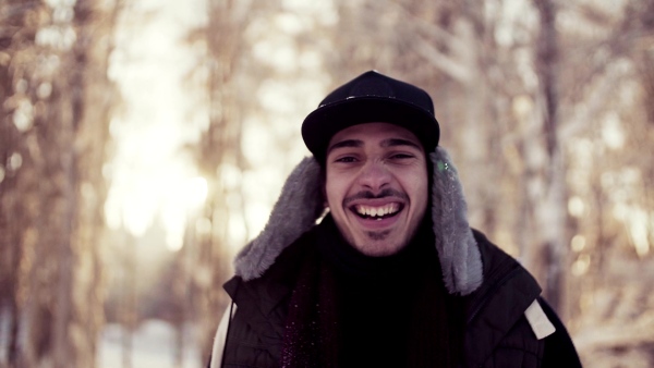 A portrait of young man standing outdoors in snow in winter forest, looking at camera. Slow motion.