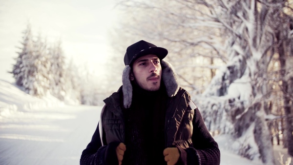 A portrait of young man standing outdoors in snow in winter forest, feeling cold. Slow motion.