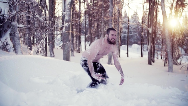 A portrait of topless young man outdoors jumping in snow in winter forest, having fun. Slow motion.