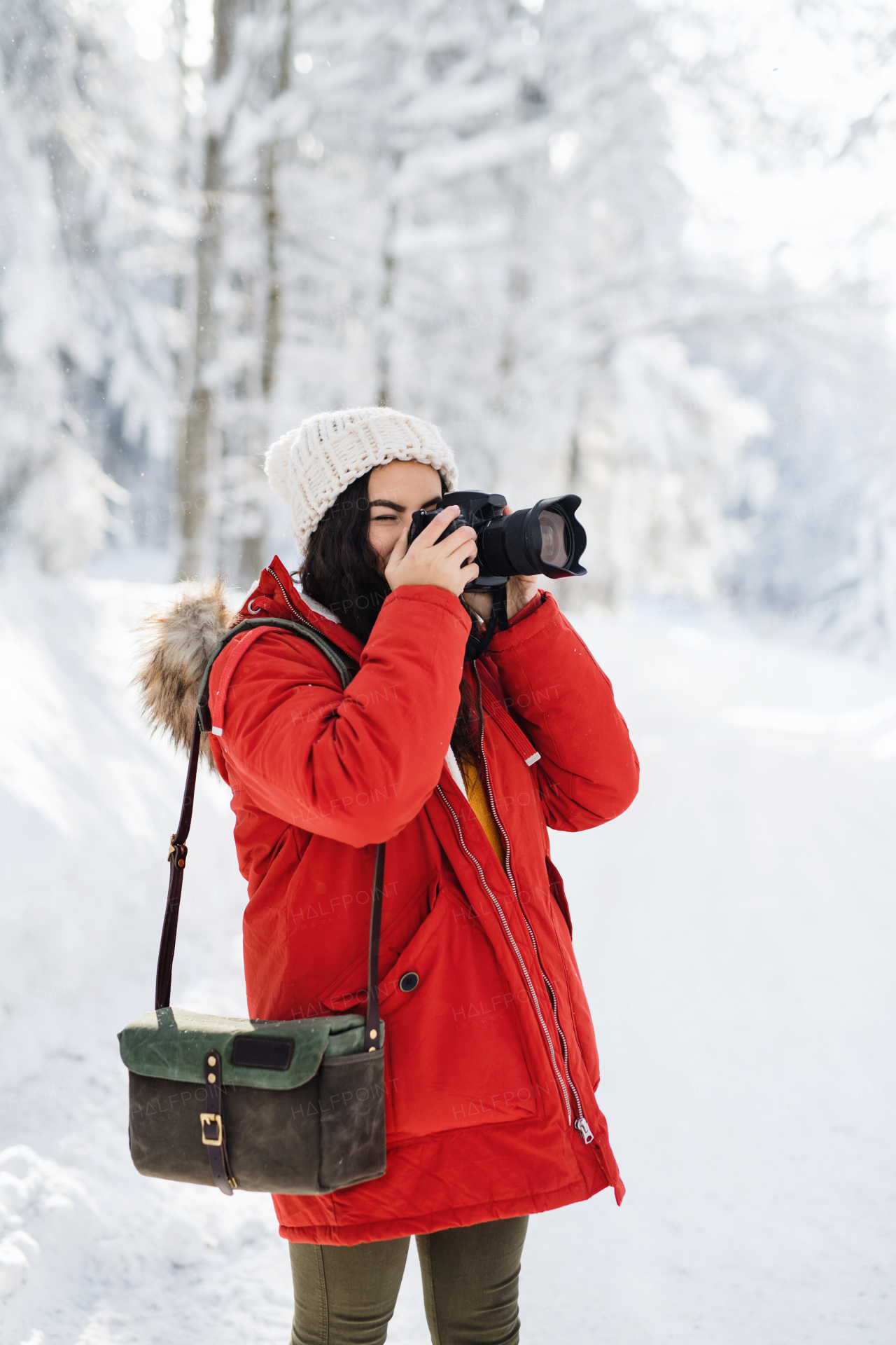 A young woman with camera standing outdoors in snow in winter forest, taking photos.