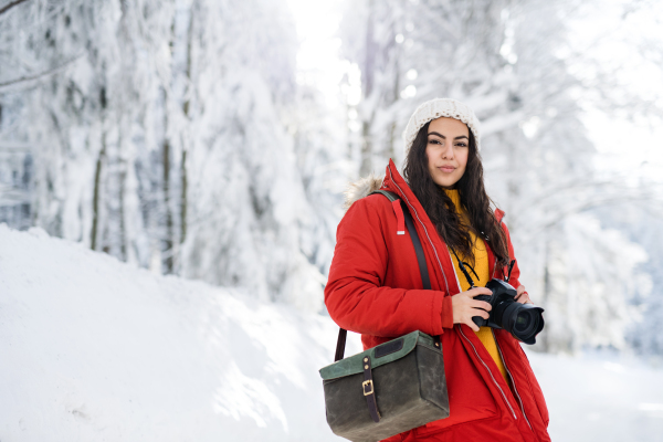 A young woman with camera standing outdoors in snow in winter forest.