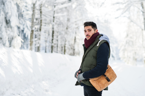 A portrait of young man standing outdoors in snow in winter forest, looking at camera.