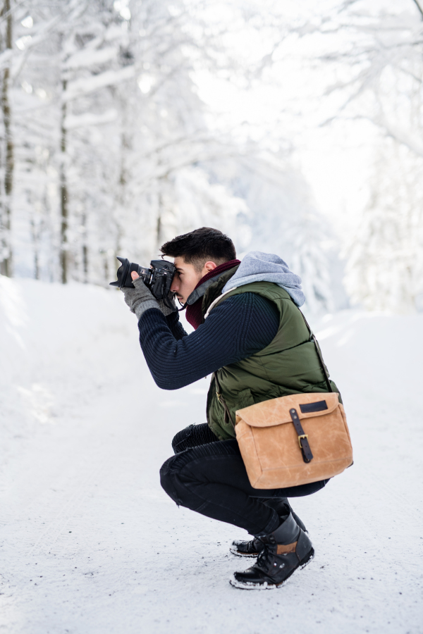 Side view of young man with camera standing outdoors in snow in winter forest, taking photos.