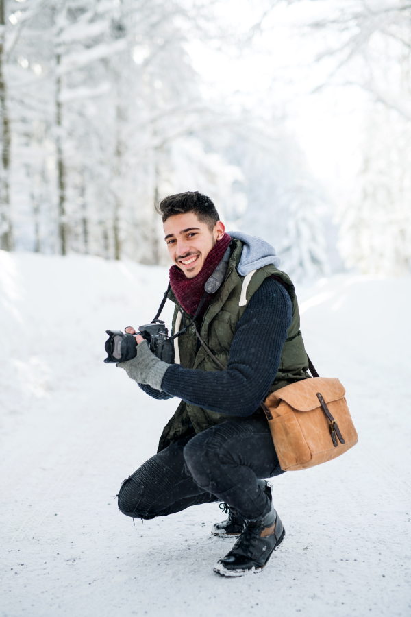 A young man with camera outdoors in snow in winter forest, looking at camera.