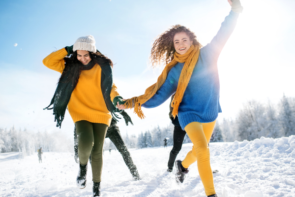 A group of young cheerful friends on a walk outdoors in snow in winter forest, holding hands and running.