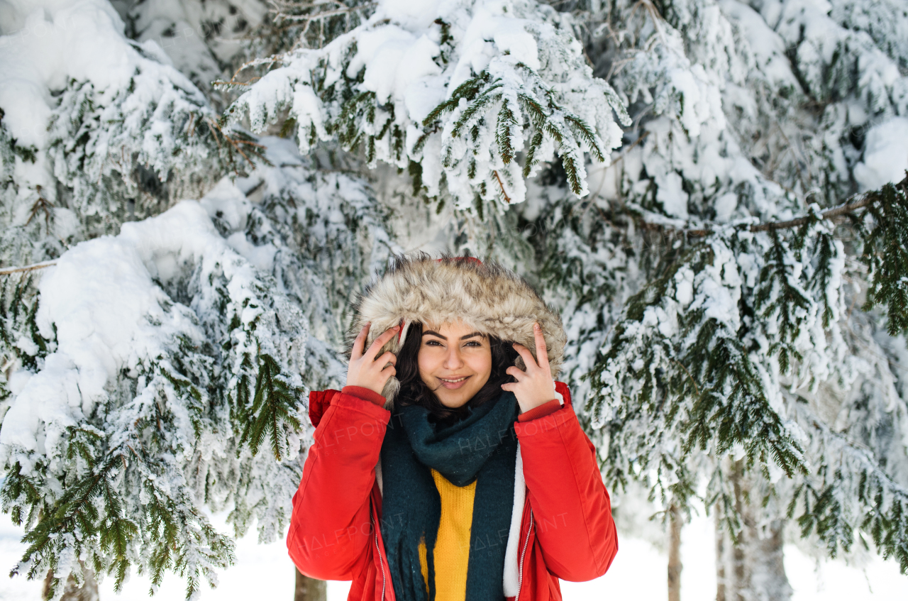 A front view portrait of young woman standing outdoors in snowy winter forest, wearing fur hood.