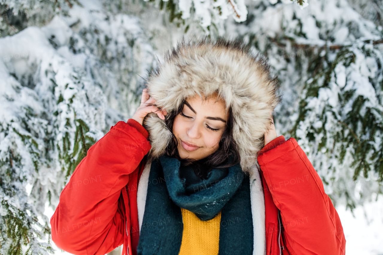 A front view portrait of young woman standing outdoors in snowy winter forest, wearing fur hood.