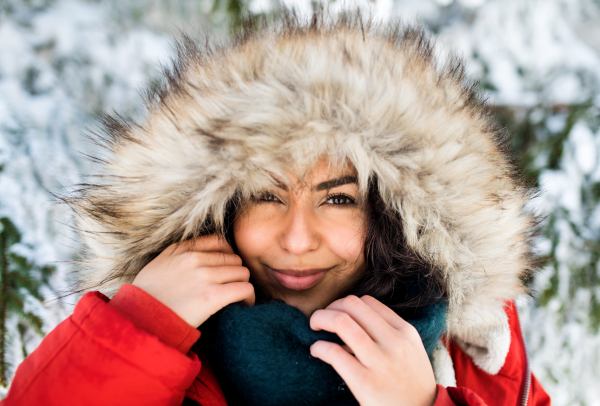 A front view portrait of young woman standing outdoors in snowy winter forest, wearing fur hood.