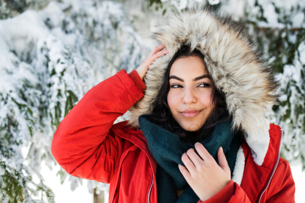 A front view portrait of young woman standing outdoors in snowy winter forest, wearing fur hood.