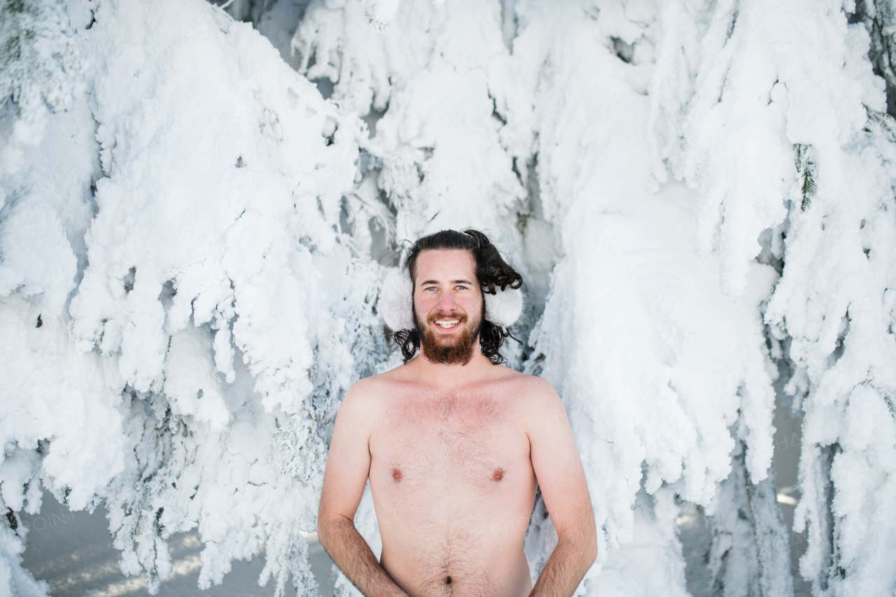 A topless young man outdoors in snow in winter forest, looking at camera.