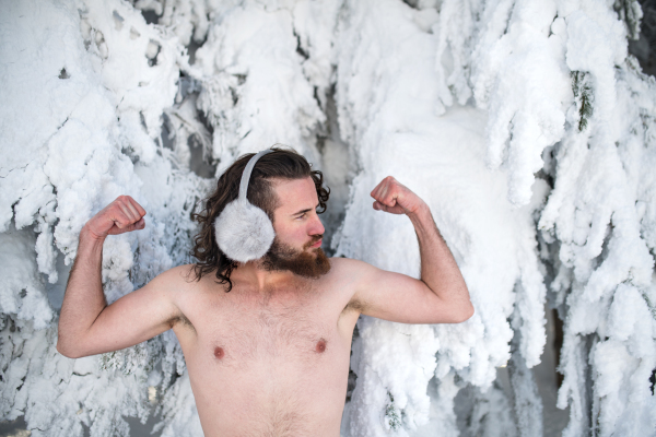 A portrait of topless young man outdoors in snow in winter forest, having fun.