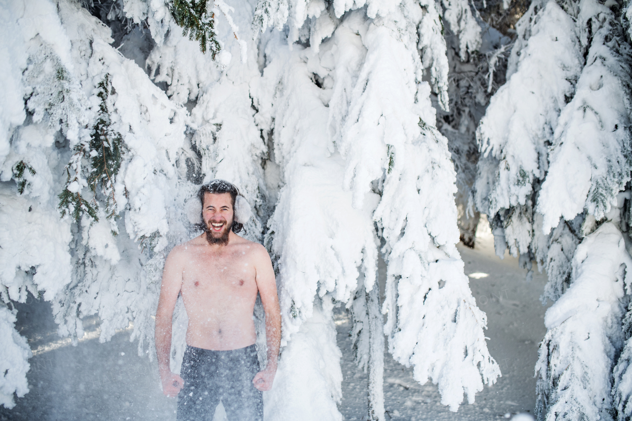 A portrait of topless young man outdoors in snow in winter forest, having fun.