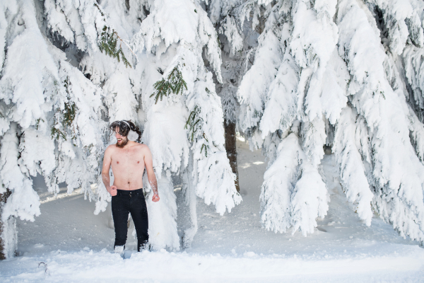 A portrait of topless young man outdoors in snow in winter forest, having fun.