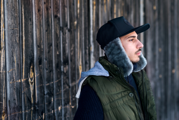 Side view portrait of young man standing against wooden background outdoors in winter.