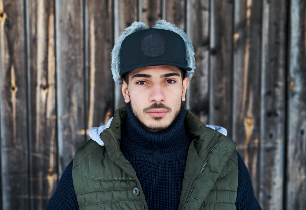 Front view portrait of young man with cap standing against wooden background outdoors in winter, close-up.