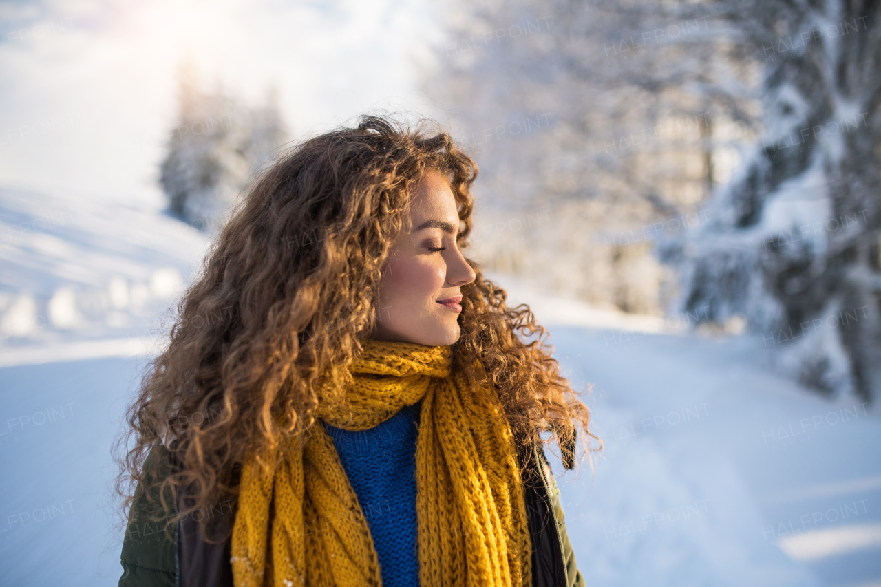 A front view portrait of young woman standing outdoors in snowy winter forest, eyes closed.