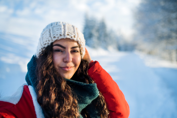 A front view portrait of young woman standing outdoors in snowy winter forest, wearing woollen hat.