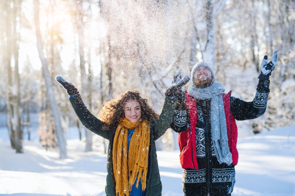 A portrait of couple standing outdoors in snow in winter forest, throwing snow.