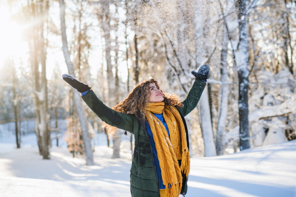 A portrait of young woman standing outdoors in snowy winter forest, throwing snow.