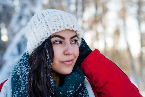 A front view portrait of young woman standing outdoors in snowy winter forest, wearing woollen hat.