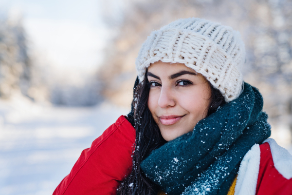 A front view portrait of young woman standing outdoors in snowy winter forest, wearing winter hat.