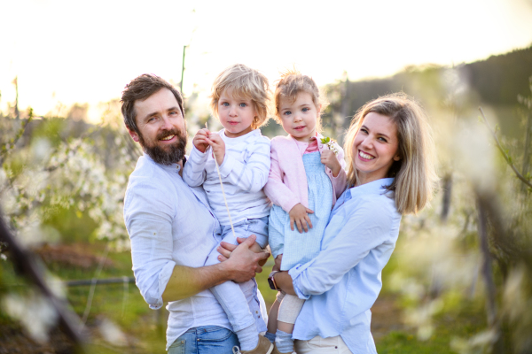 Front view of family with two small children standing outdoors in orchard in spring, looking at camera.