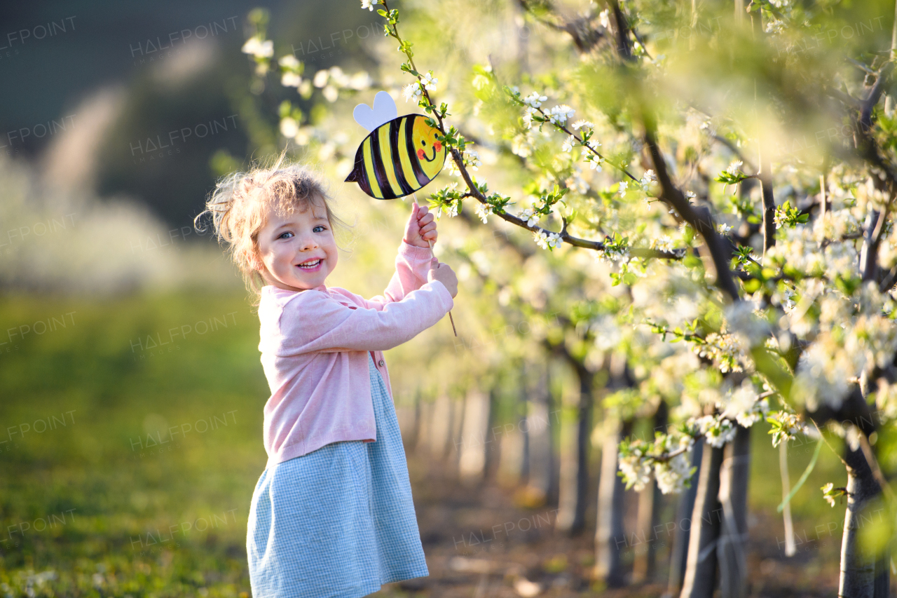 Side view of small toddler girl standing outdoors in orchard in spring, holding paper bee.