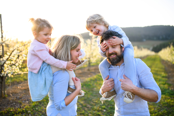 Front view of family with two small children standing outdoors in orchard in spring, piggyback ride.