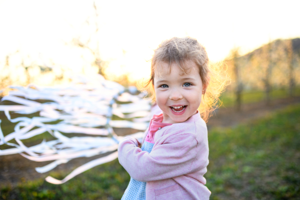 Cheerful small toddler girl standing outdoors in orchard in spring, holding hand ribbon ring.