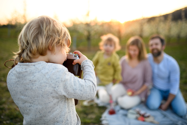 Small boy with camera taking photograph of family on picnic outdoors in spring nature at sunset.