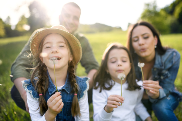 Front view of happy family with two small daughters sitting outdoors in spring nature, blowing dandelion seeds.