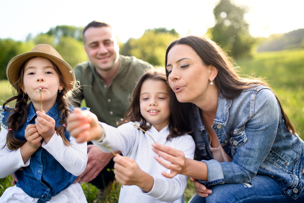 Front view of happy family with two small daughters sitting outdoors in spring nature, blowing dandelion seeds.