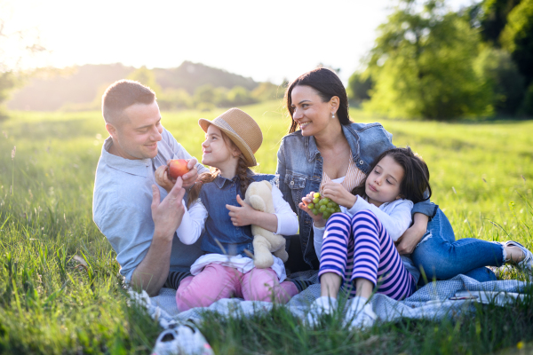 Front view of happy family with two small daughters sitting outdoors in spring nature, having picnic.