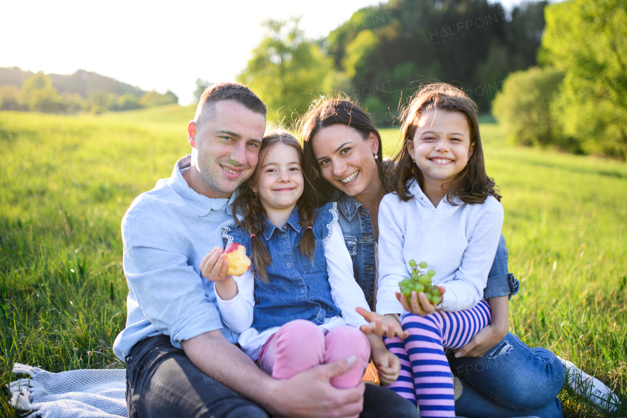 Front view of happy family with two small daughters sitting outdoors in spring nature, having picnic.