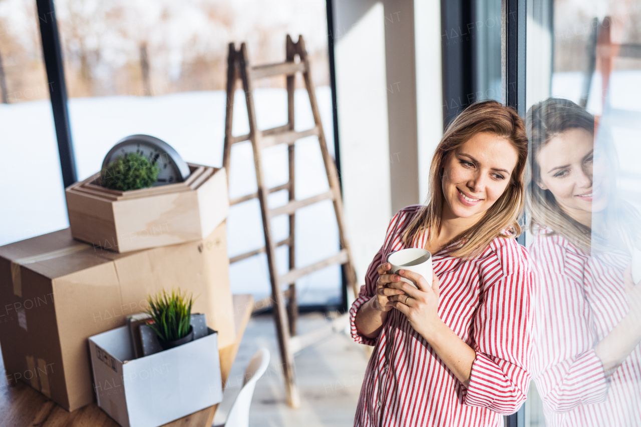 A happy young woman moving in new home, leaning on a window and drinking coffee.