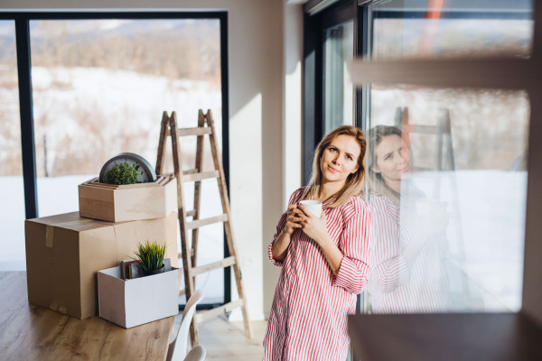 A happy young woman moving in new home, leaning on a window and drinking coffee.