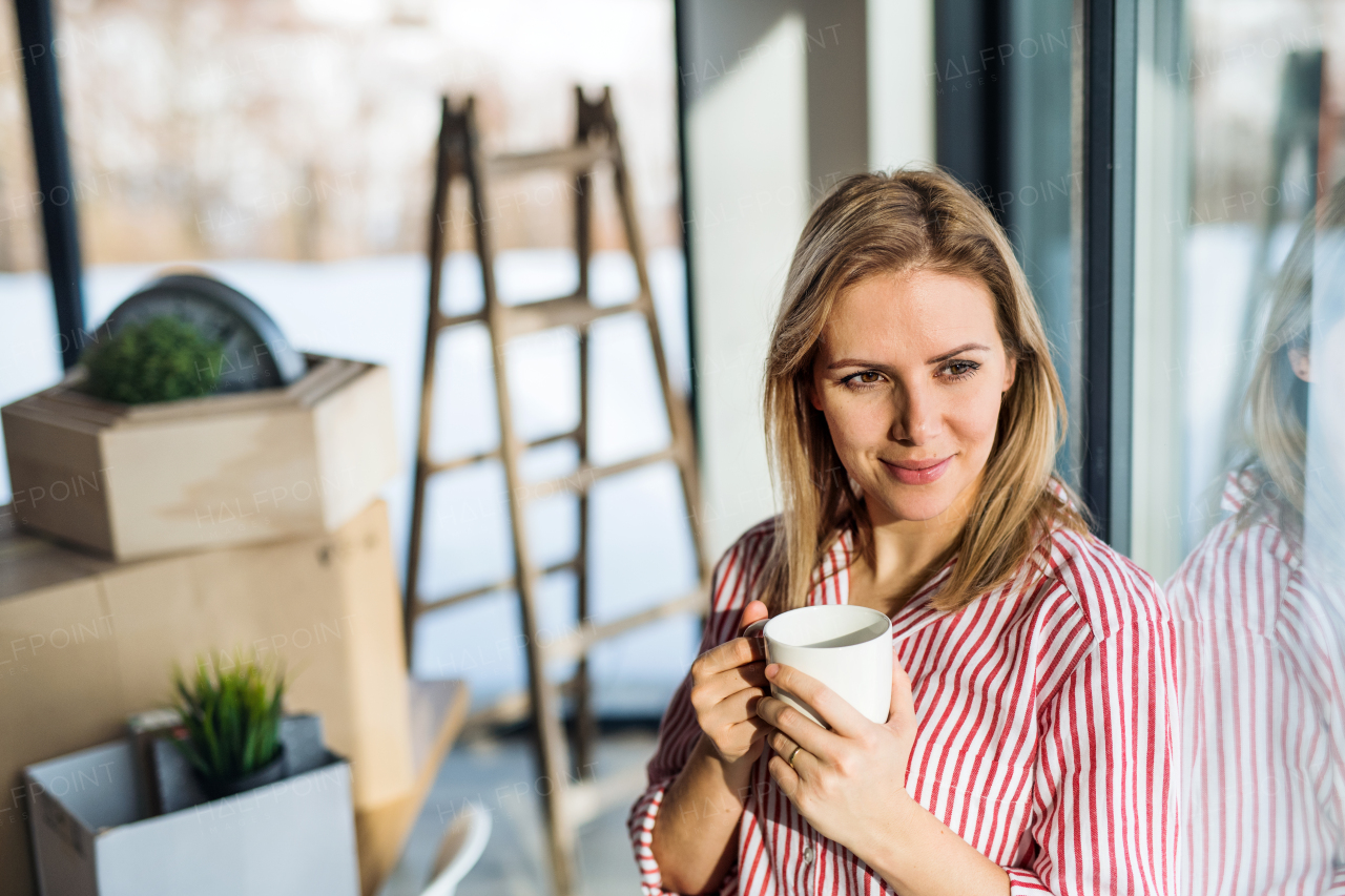 A happy young woman moving in new home, leaning on a window and drinking coffee.