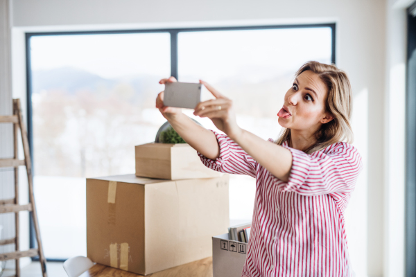 A happy young woman with smartphone moving in new home, grimacing when taking selfie.