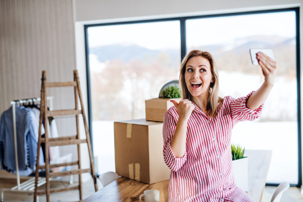 A happy young woman with smartphone moving in new home, grimacing when taking selfie.