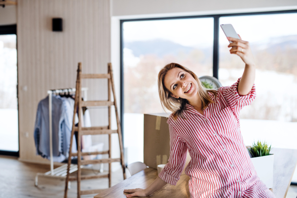 A happy young woman with smartphone moving in new home, taking selfie.