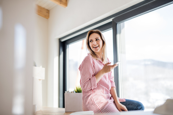 A happy young woman with smartphone moving in new home, sitting on table.