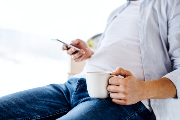 A midsection of unrecognizable man with smartphone and coffee sitting indoors, text messaging.