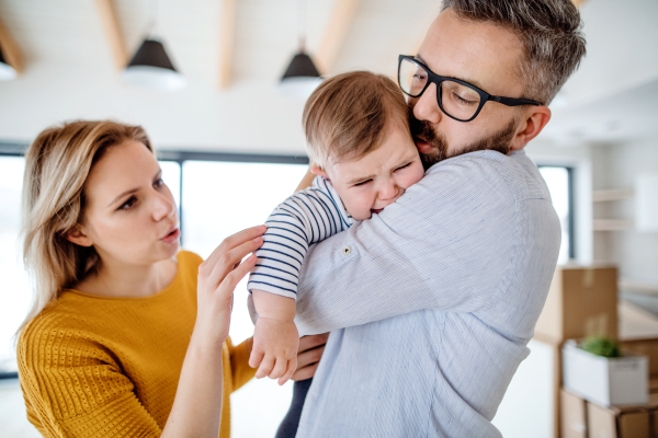 A young family with an unhappy crying toddler girl standing indoors at home.