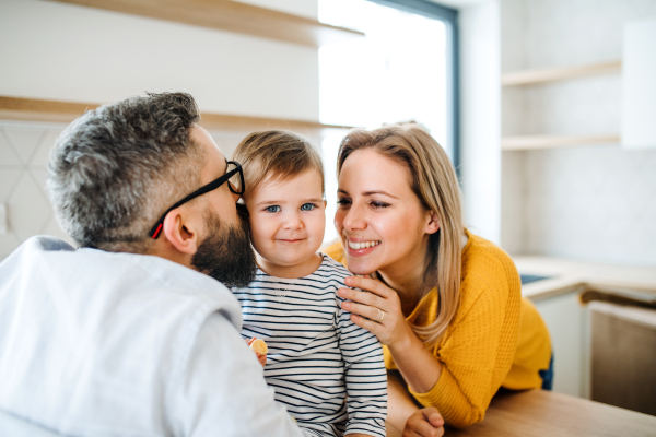 A portrait of young happy family with a toddler girl indoors in kitchen, kissing.