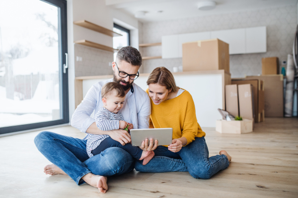 A young family with a toddler girl sitting barefooton the floor when moving in new home, using tablet.