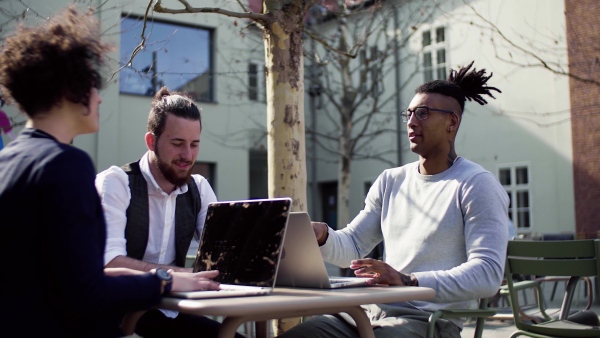 A group of young businesspeople with laptop in outdoor cafe, talking. A start-up concept.