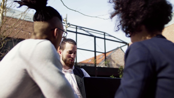 A group of young businesspeople with laptop in outdoor cafe, talking. A start-up concept.