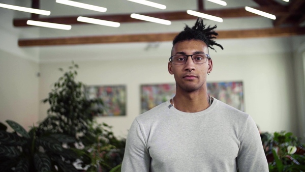 A portrait of young mixed race businessman with dreadlocks in office, looking at camera. Slow motion.