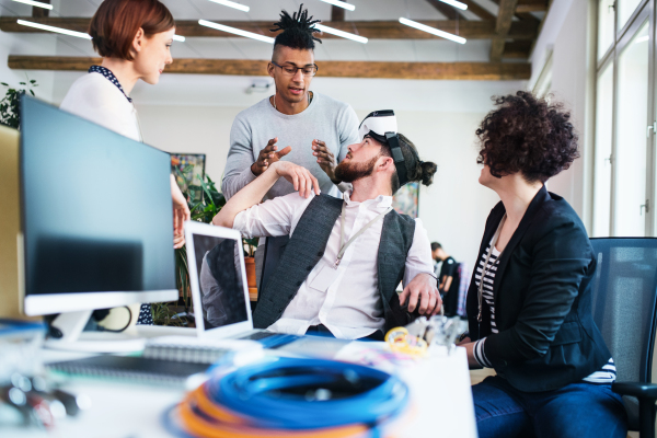 A group of young businesspeople with VR goggles talking in office, start-up concept.