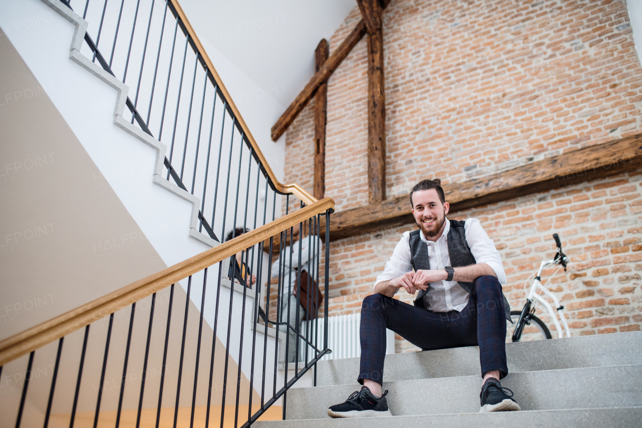 A young businessman sitting on stairs in office building, looking at camera.